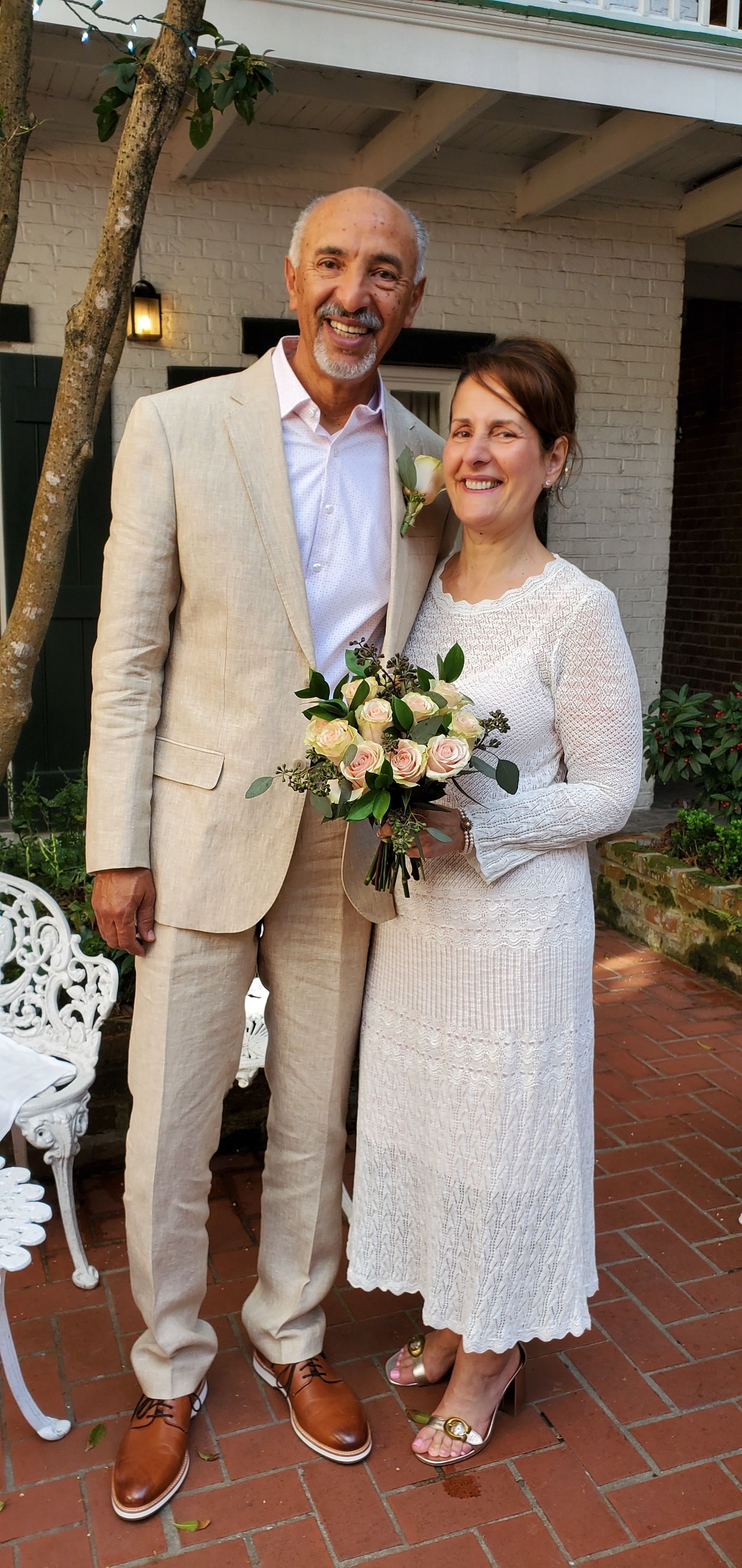 Couple posing outdoors at a wedding, holding a bouquet, in white formal attire