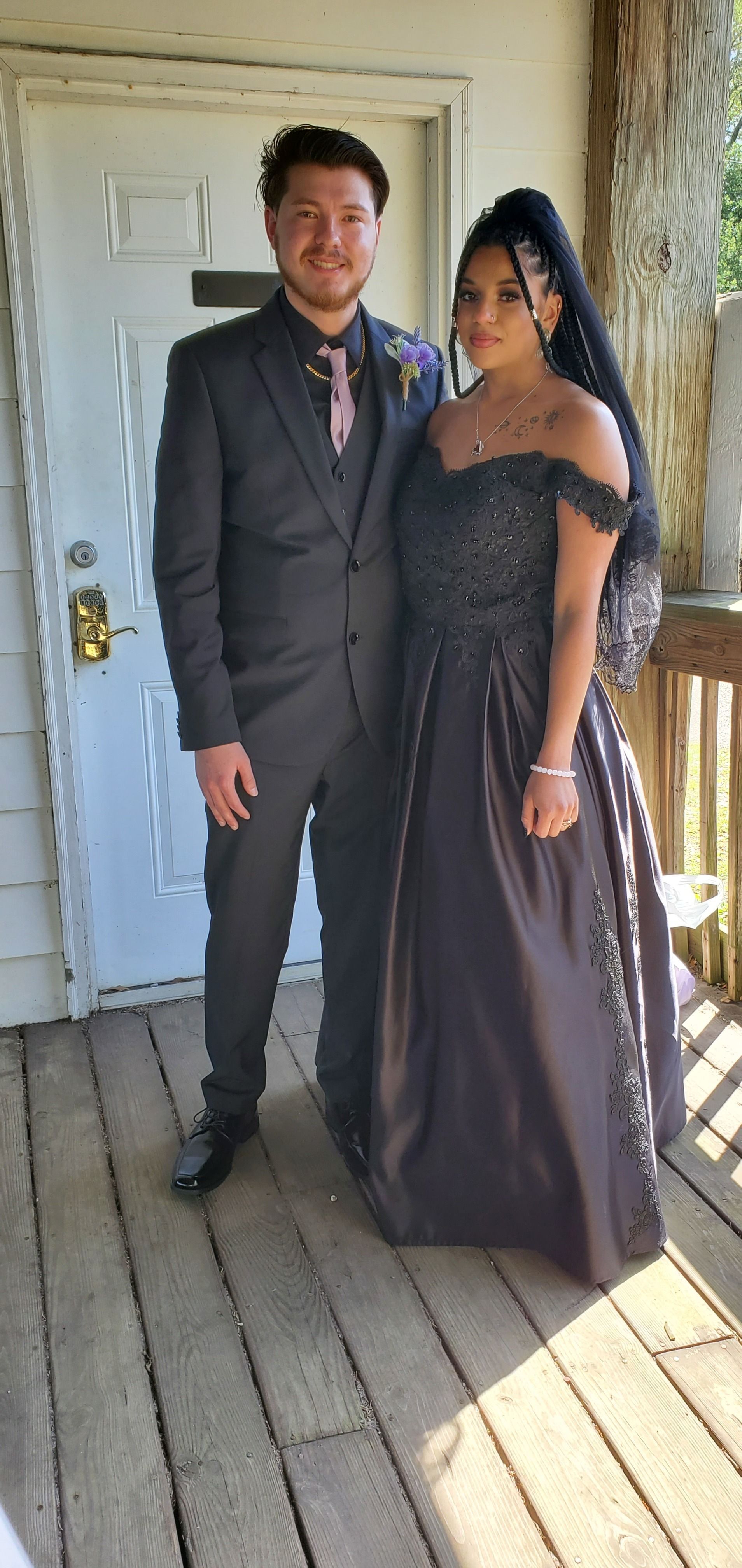 Two people in formal attire standing on a porch, smiling beside a white door and railing.