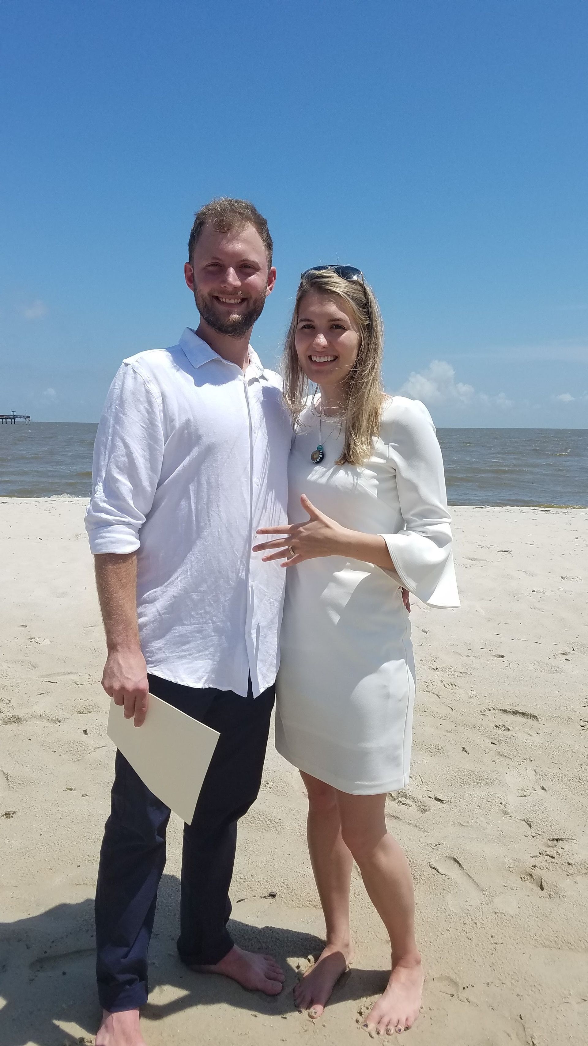 Couple standing on a sunny beach, posing in white outfits with the ocean behind them.