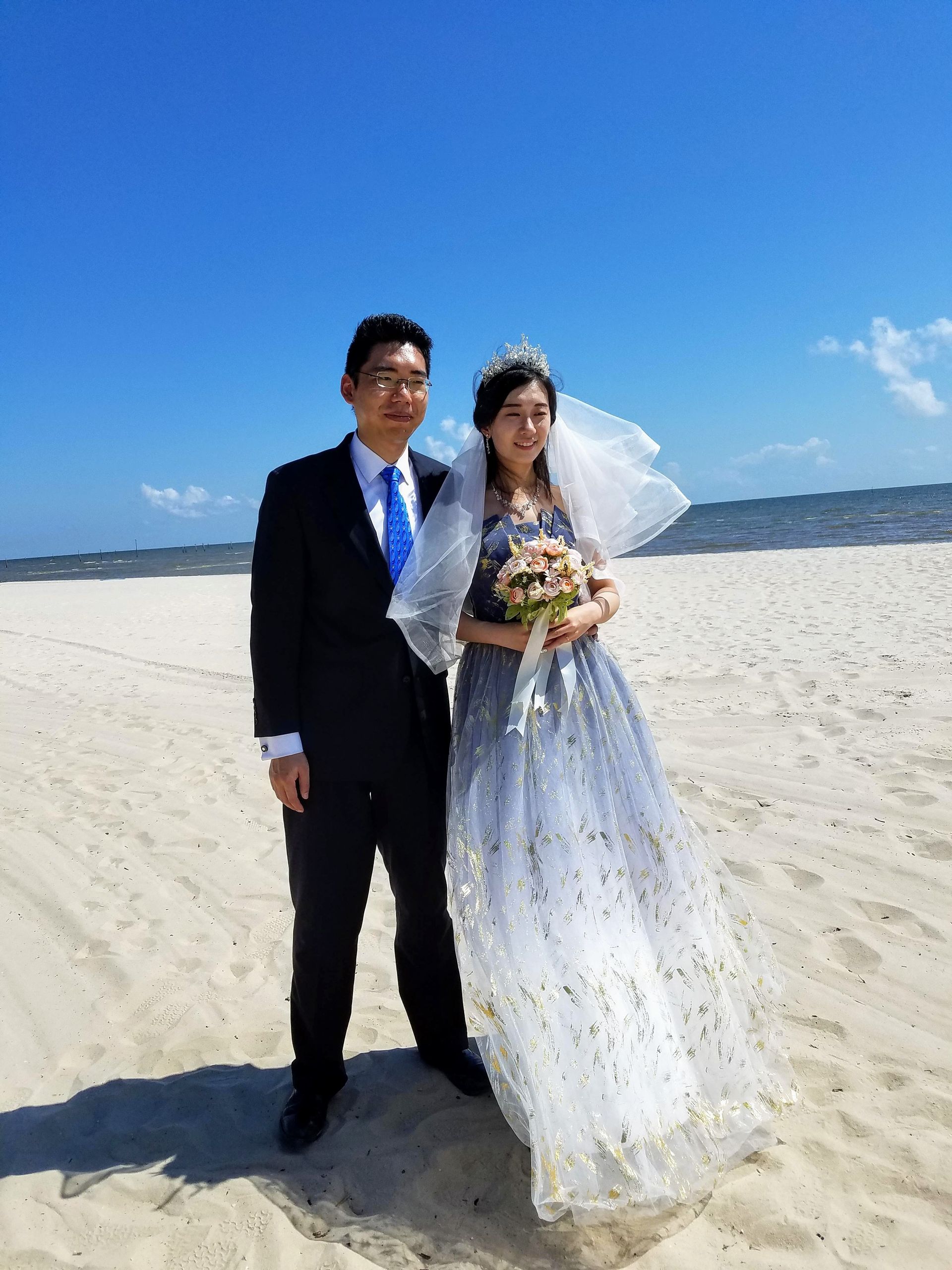 Wedding couple standing on a sunny beach, bride in white gown and groom in black suit holding bouquet.