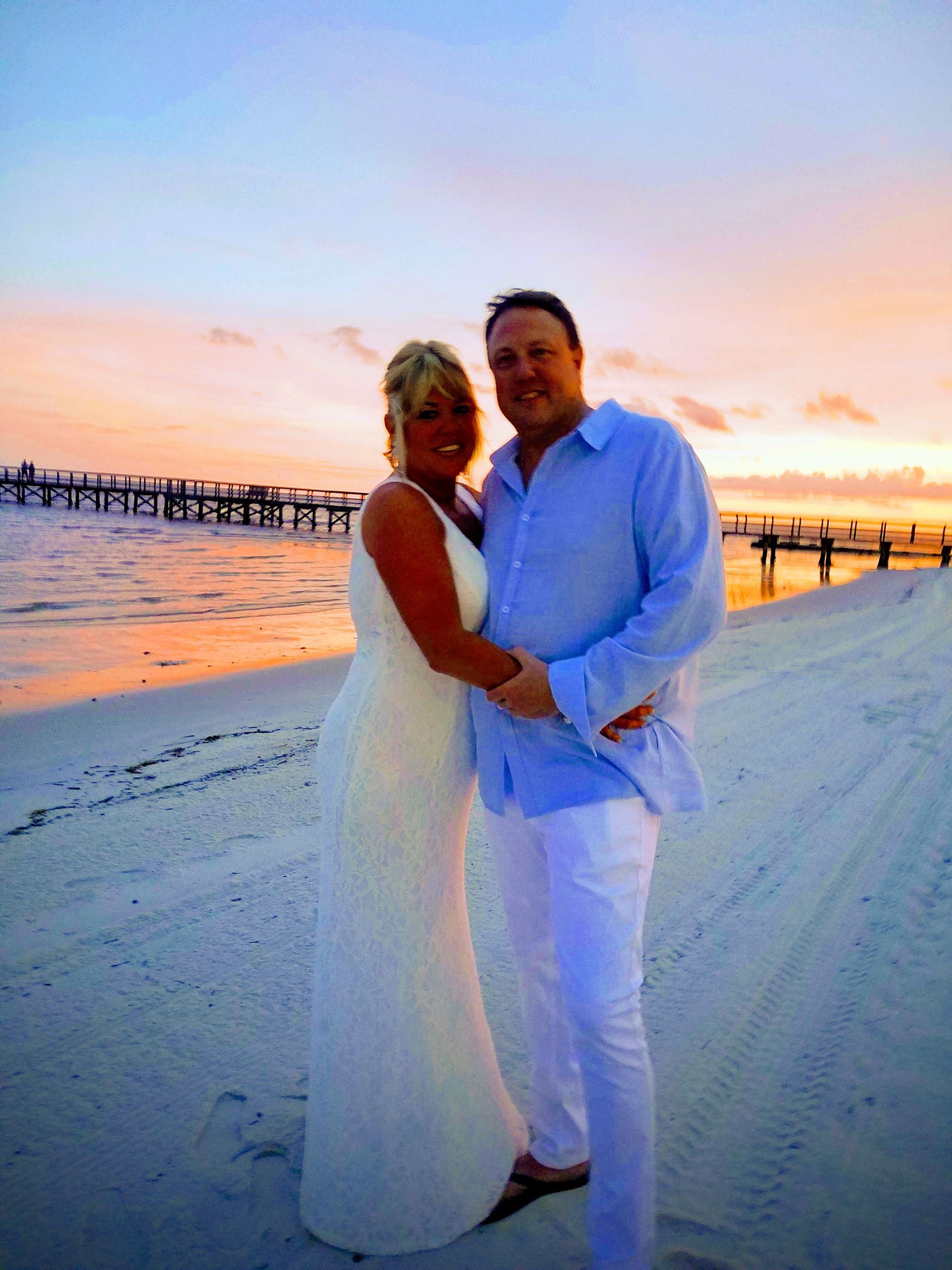 Couple in white on a beach at sunset, standing hand in hand near the shoreline.
