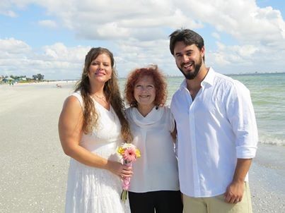 Three people smiling on a sunny beach, with waves and a cloudy sky behind them.
