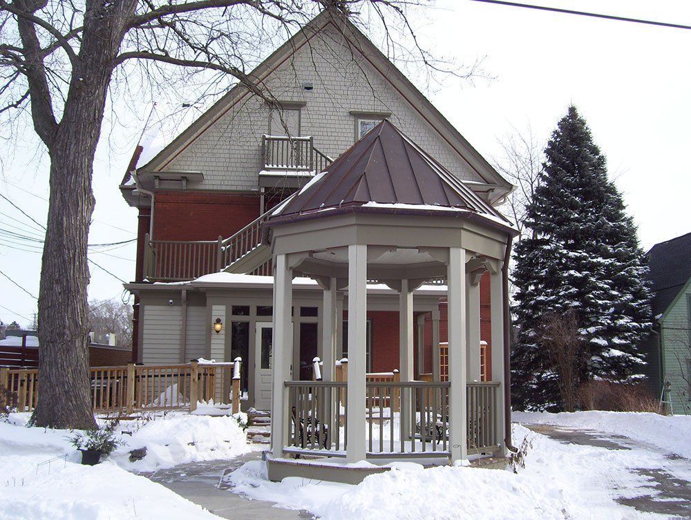 Snowy yard with a red house and a white gazebo in front of a large evergreen tree.