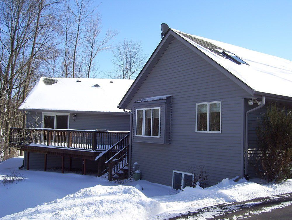 Snow-covered gray house with deck and bare trees in a winter yard