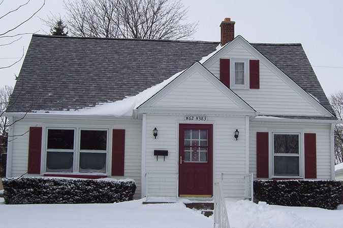 Snow-covered white house with red door and shutters, viewed from the front.