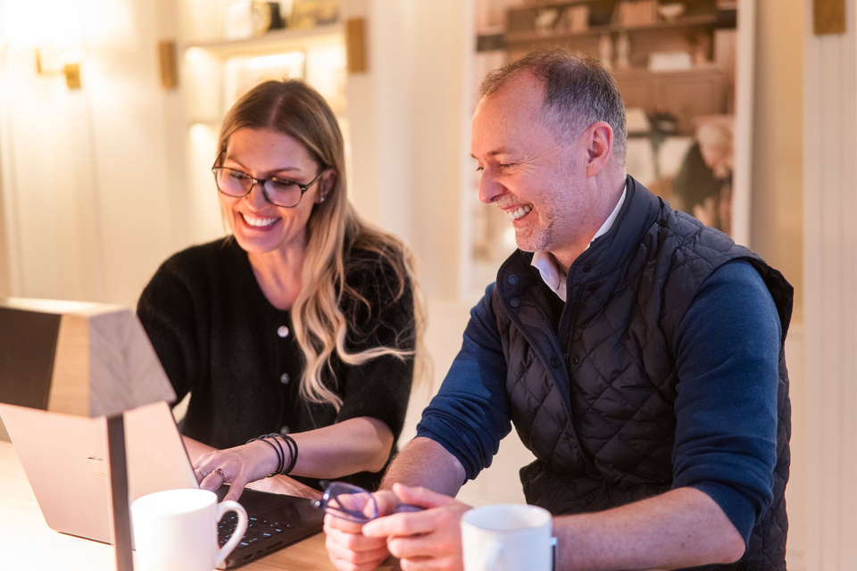 Two people smiling and working at a laptop in a cozy, warmly lit room