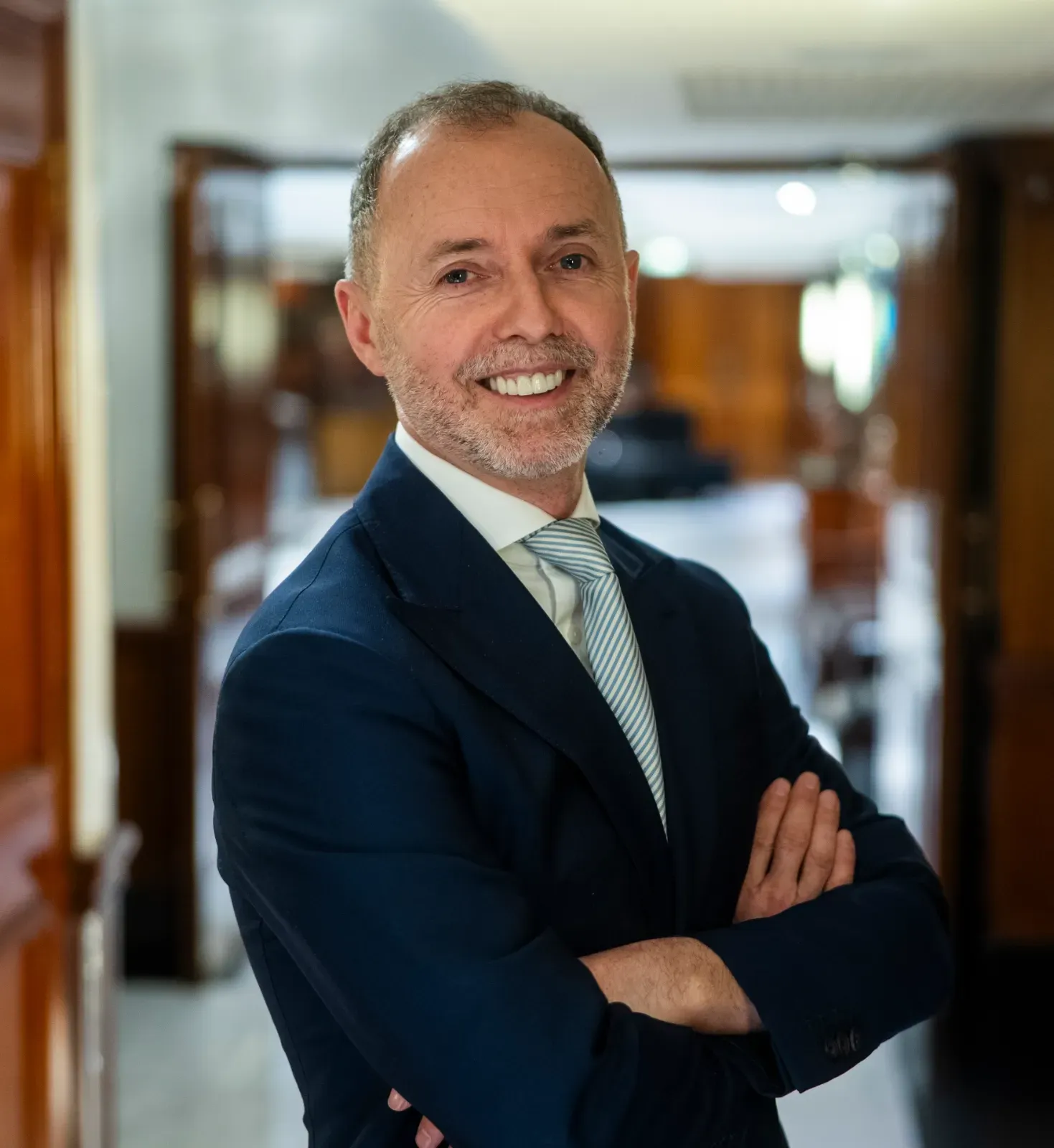 Smiling businessman in a navy suit with arms crossed in a blurred office hallway.