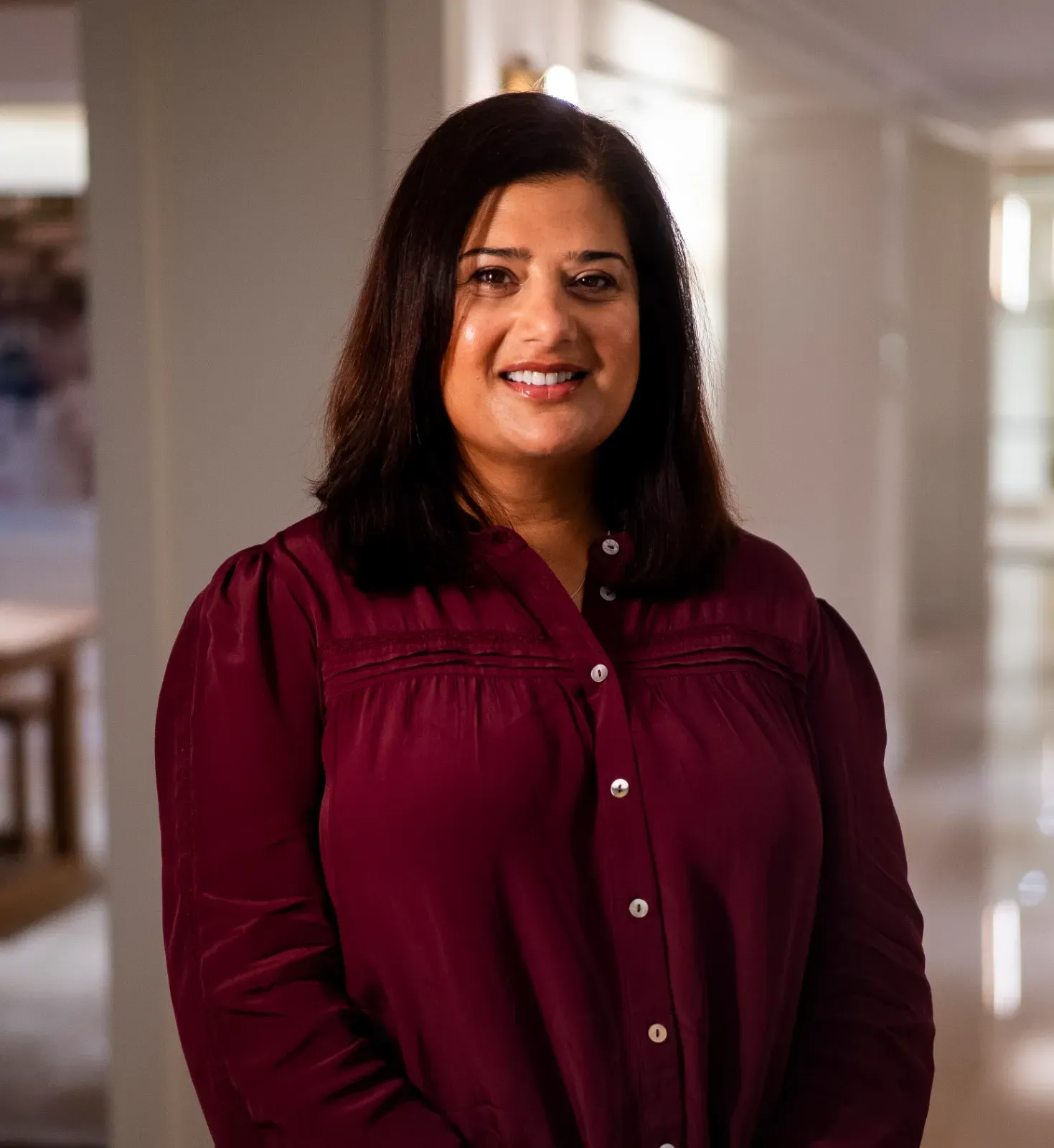 Woman in a burgundy blouse standing in a bright hallway, smiling at the camera