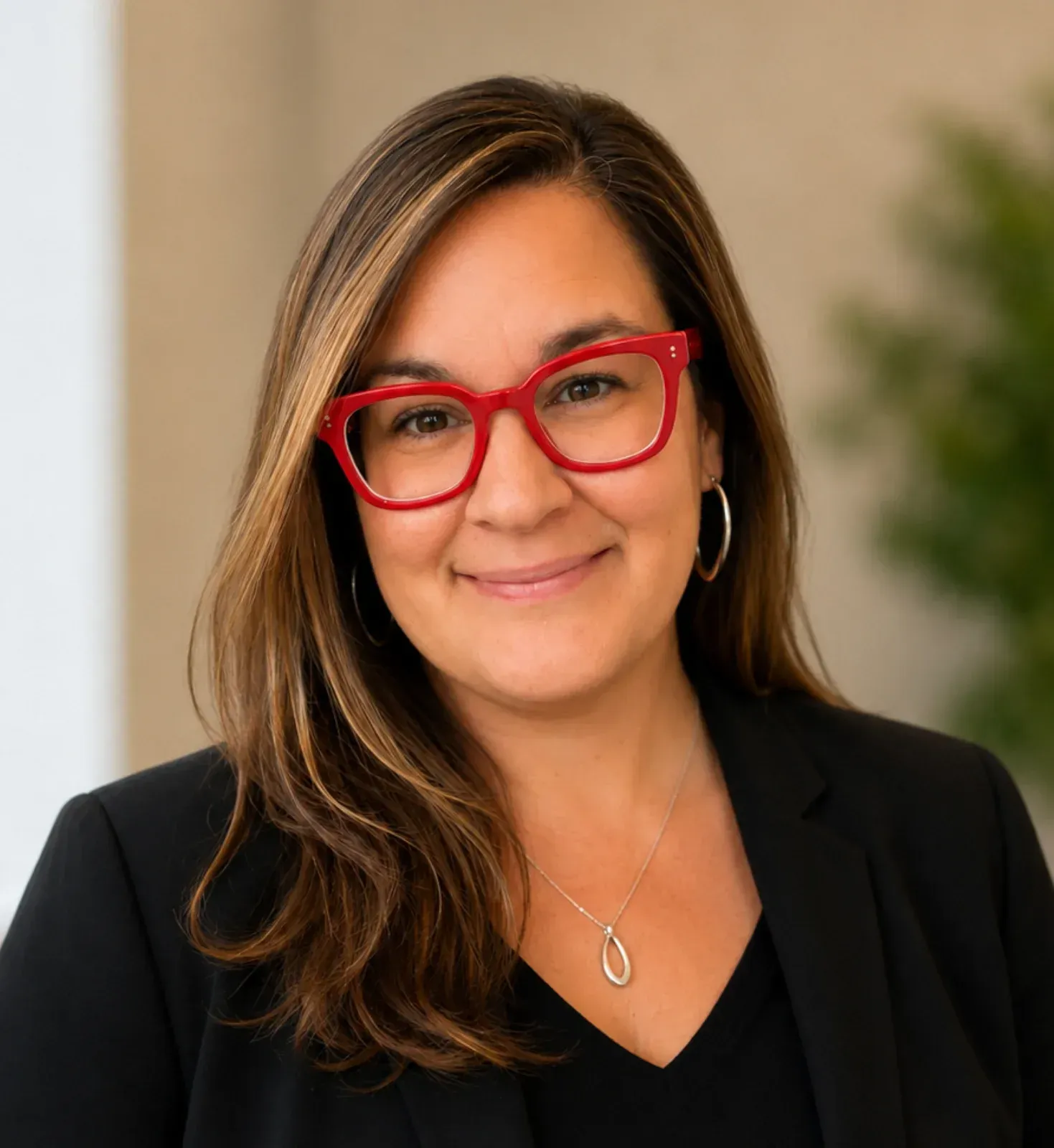 Portrait of a smiling woman with red glasses and brown hair in a black blazer against a soft background