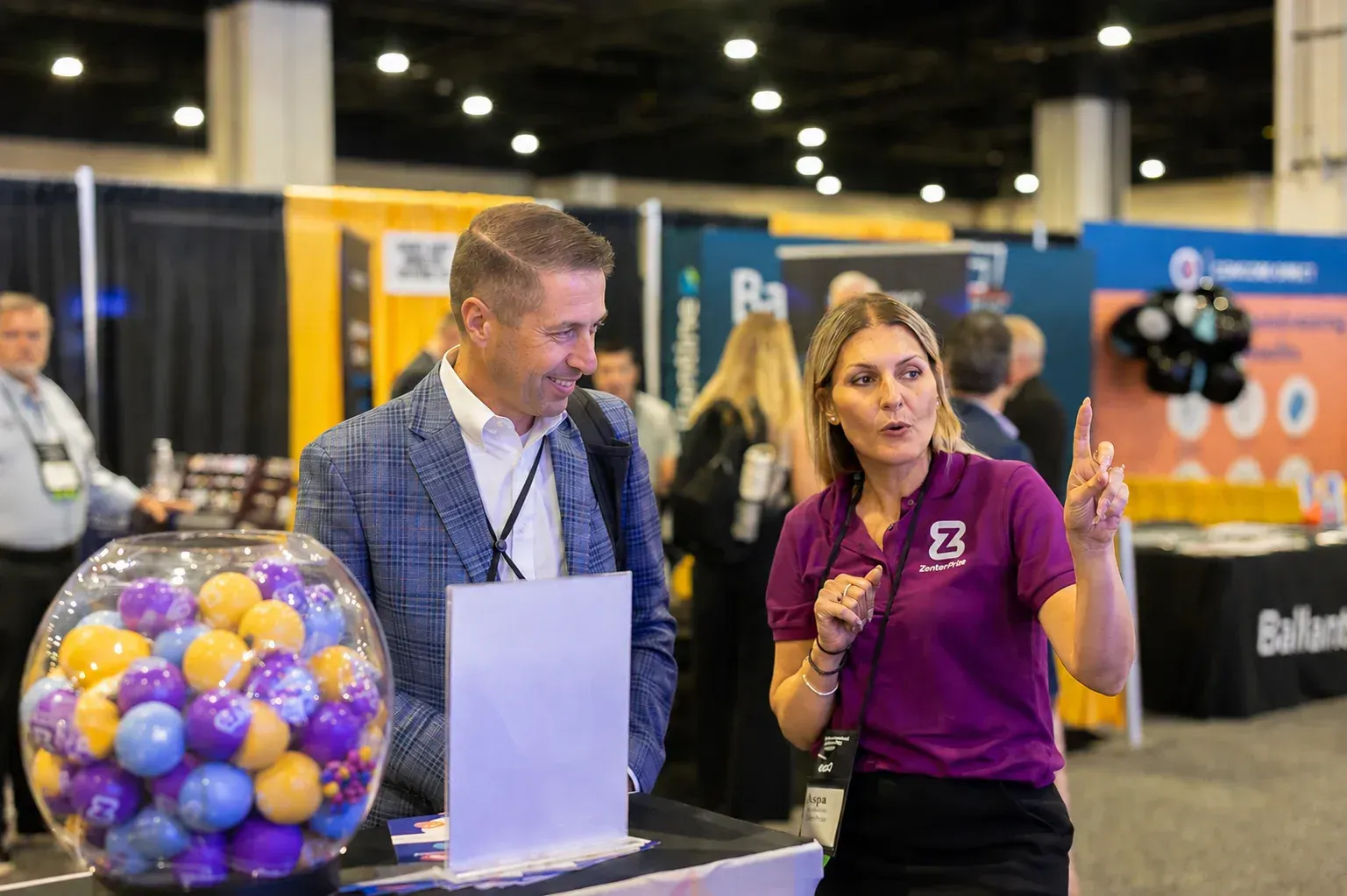 Two people at a trade show booth beside a bowl of colorful ping-pong balls