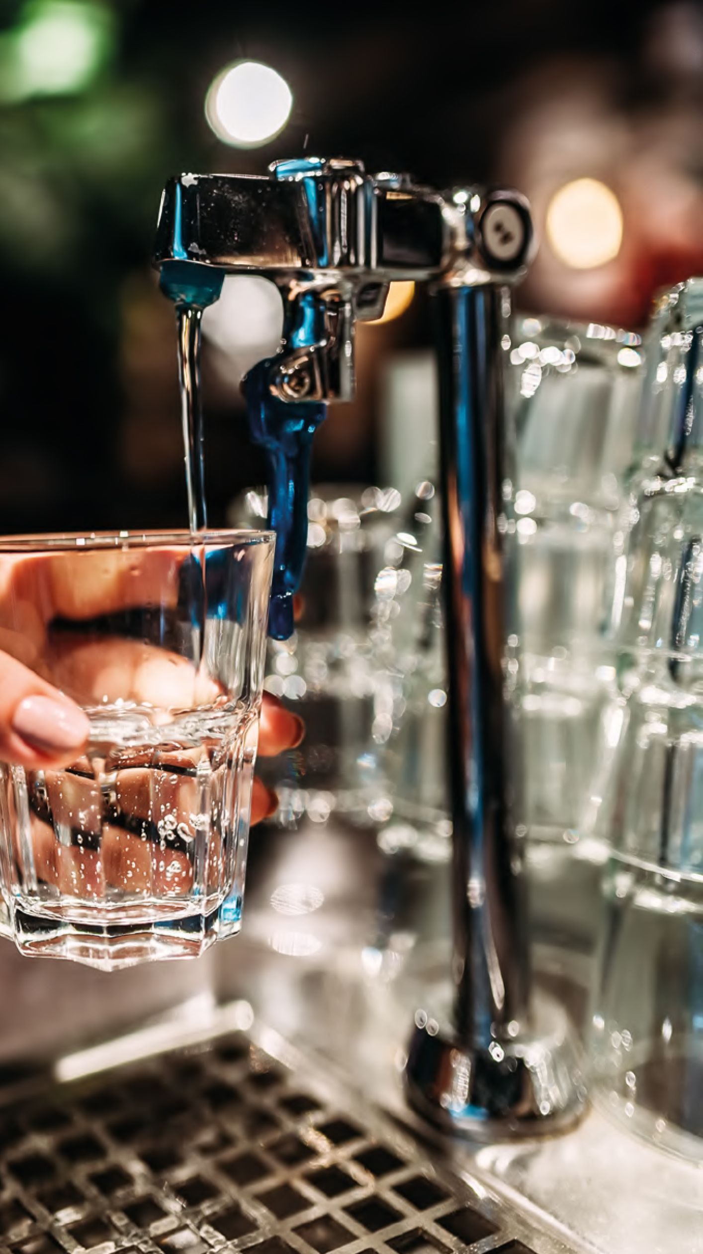 Riempire a mano un bicchiere d'acqua da un rubinetto cromato da bar, con altri bicchieri nelle vicinanze.