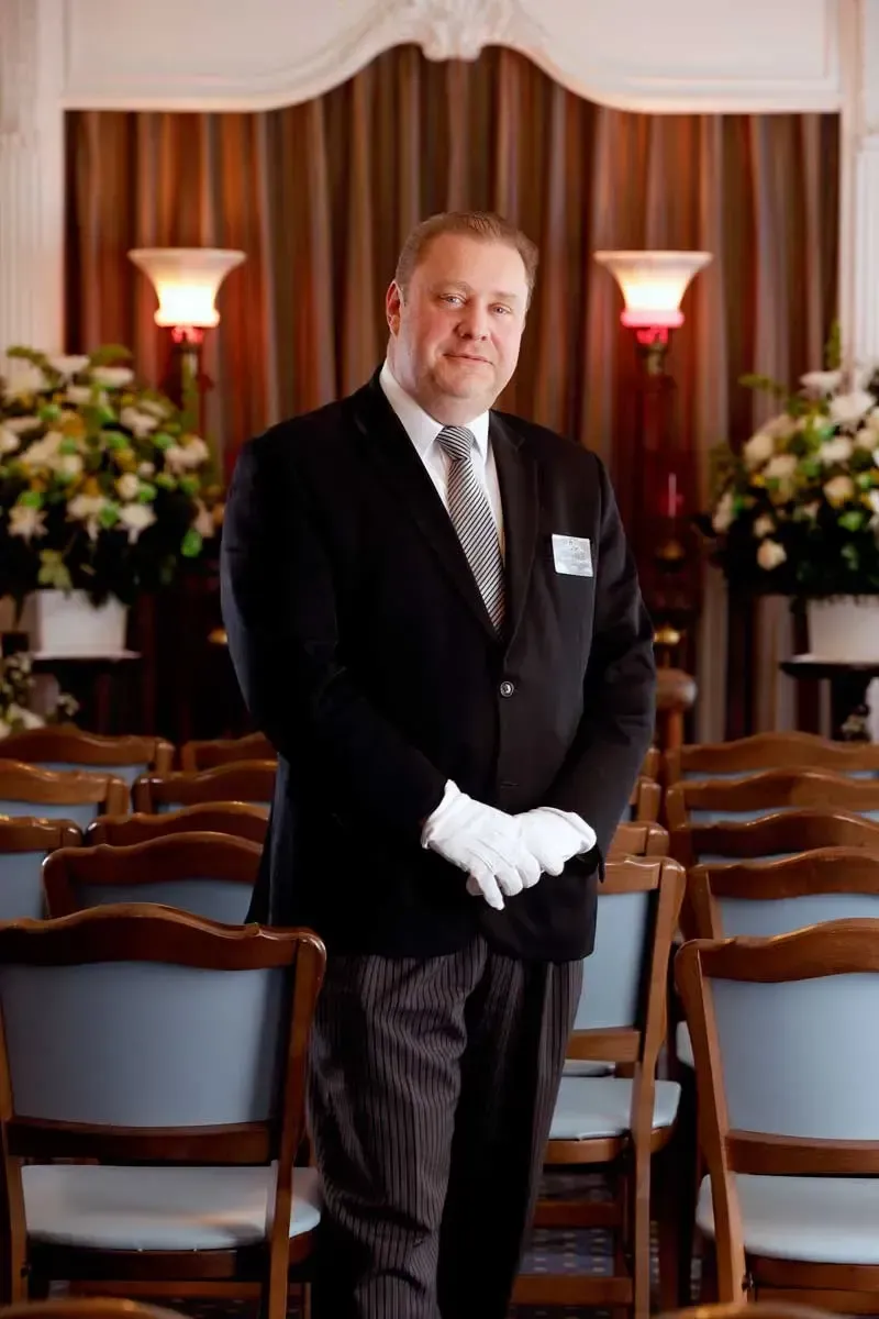 Man in a black suit with white gloves standing in a formal banquet hall with rows of chairs and flower arrangements