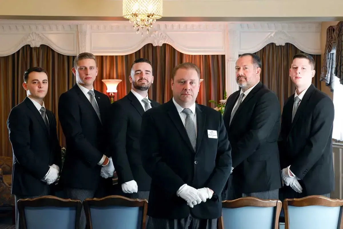Six men in black suits and white gloves stand in a formal dining room.