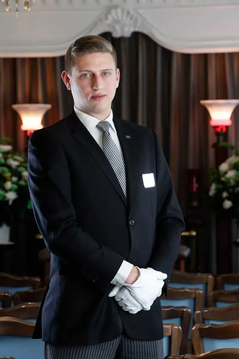 Man in a dark suit with white gloves standing in a formal event hall with rows of chairs and floral arrangements