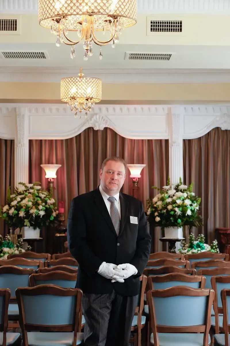 Man in a black tuxedo and white gloves standing in a formal dining room with chandeliers and floral arrangements