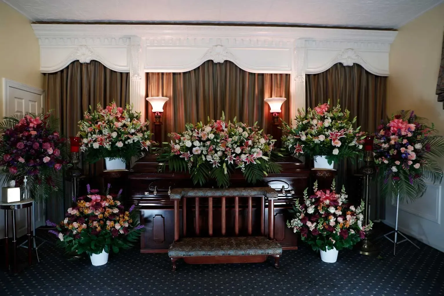 Indoor memorial altar with large pink and white floral arrangements and candles on a dark carpeted stage