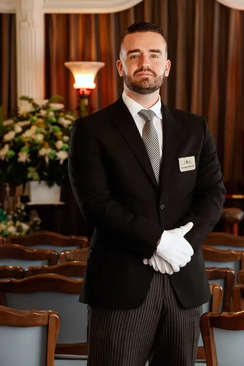 Uniformed hotel staff member standing in a formal dining room, wearing a black suit and white gloves.