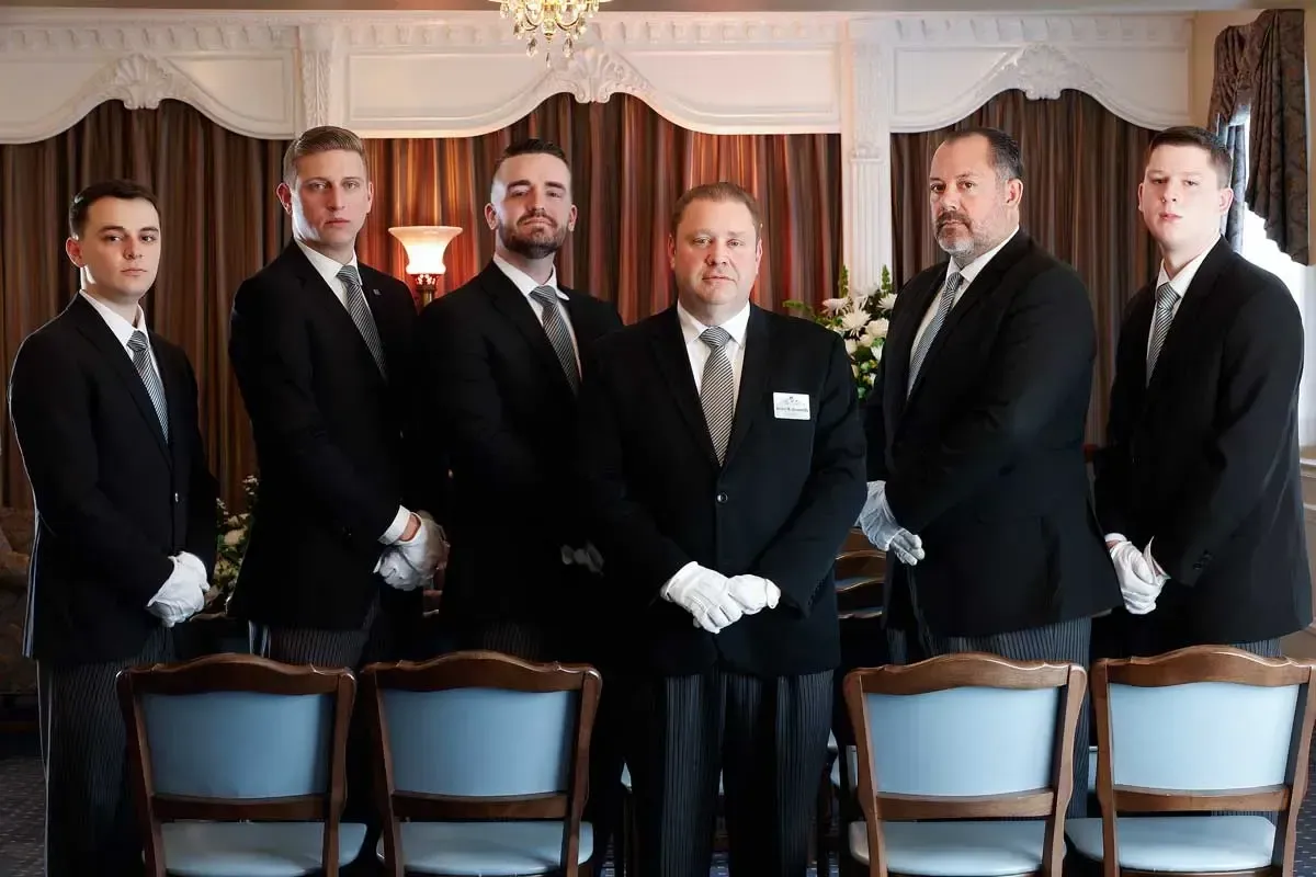 Six men in black suits stand behind chairs in a formal room, facing the camera.