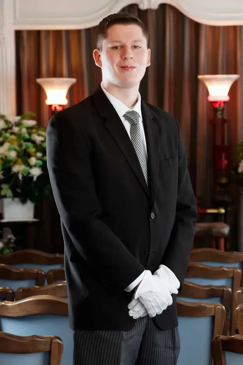 Man in a black suit with white gloves standing in a formal room with wooden chairs and lamps