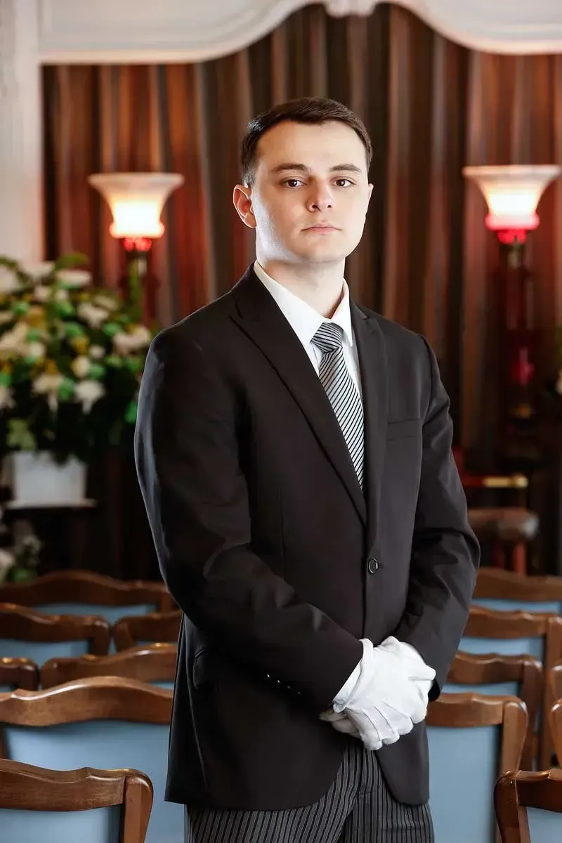 Man in a black suit and white gloves standing in a formal room with wood paneling and lamps.