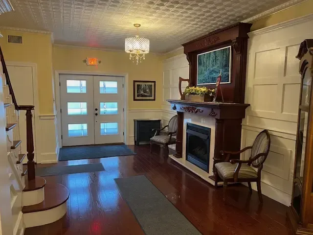 Bright foyer with double front doors, fireplace, chair, staircase, and polished hardwood floor.