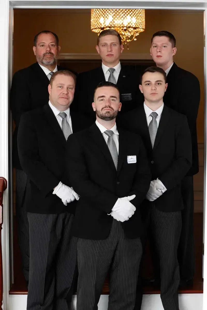Six men in black suits pose in a formal portrait indoors under a chandelier.