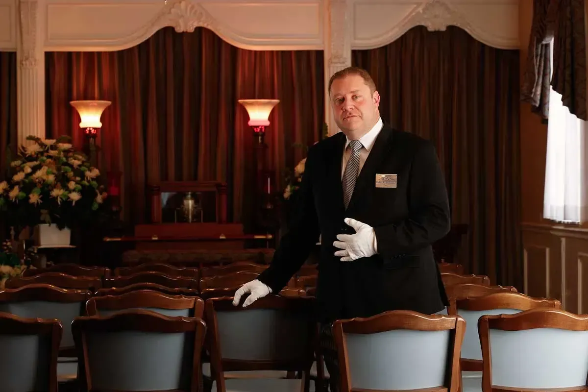 Man in black suit and white gloves standing in a dim, elegant dining room with set tables and lamps