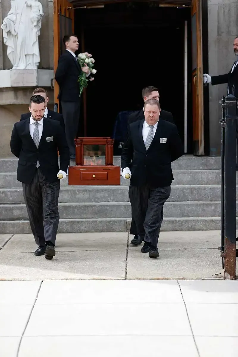Two men in dark suits walk out of a church carrying a floral wreath by a casket inside.