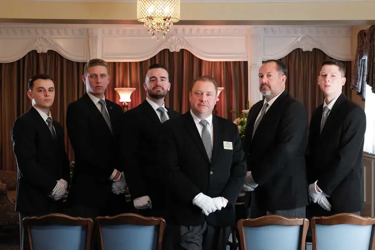 Five formally dressed men standing indoors in a chandelier-lit room, with chairs in the foreground