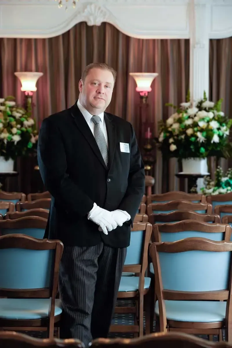 Formal staff member in black suit and white gloves standing in a banquet room with rows of chairs and flower arrangements