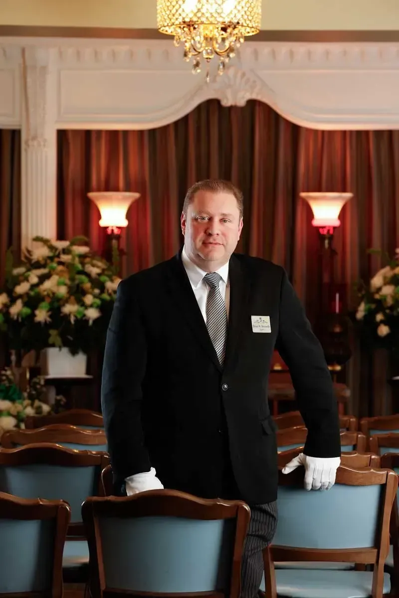 Man in a black suit standing in a formal dining room with chandeliers and dark curtains