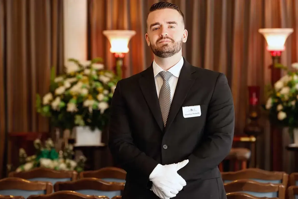 Man in black suit and white gloves standing in a formal dining room with floral arrangements and warm lighting