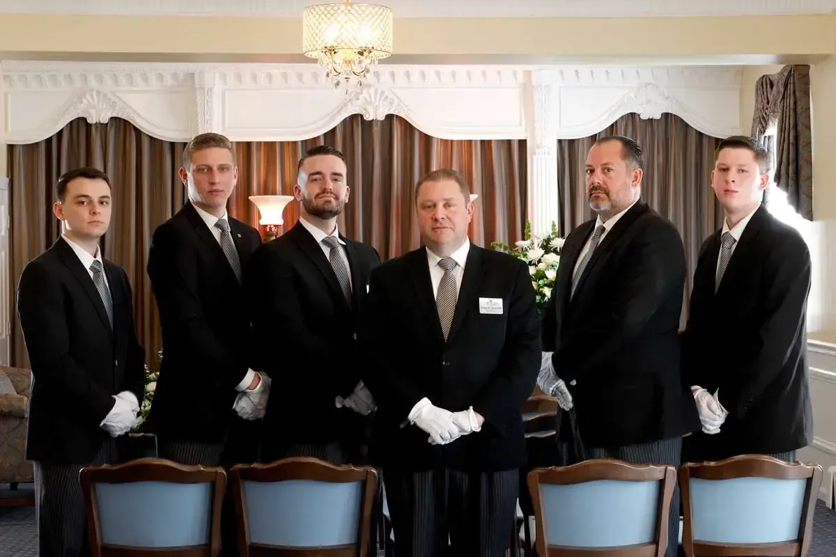Six men in black suits and white gloves stand in a formal dining room.