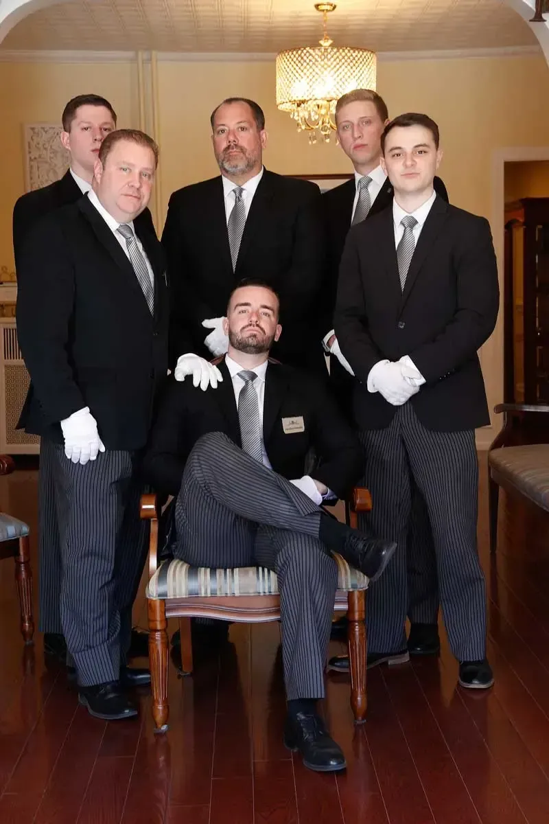 Five men in dark suits and white gloves pose indoors beneath a chandelier.