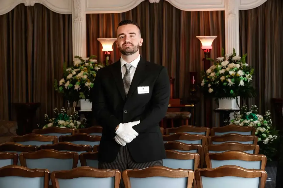 Formal usher in black suit and white gloves standing in a decorated venue before rows of chairs