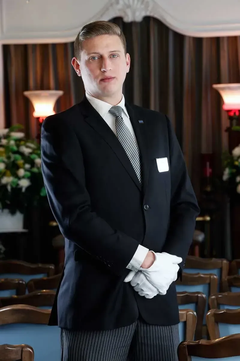 Man in a dark suit and white gloves standing indoors between rows of chairs in a formal room