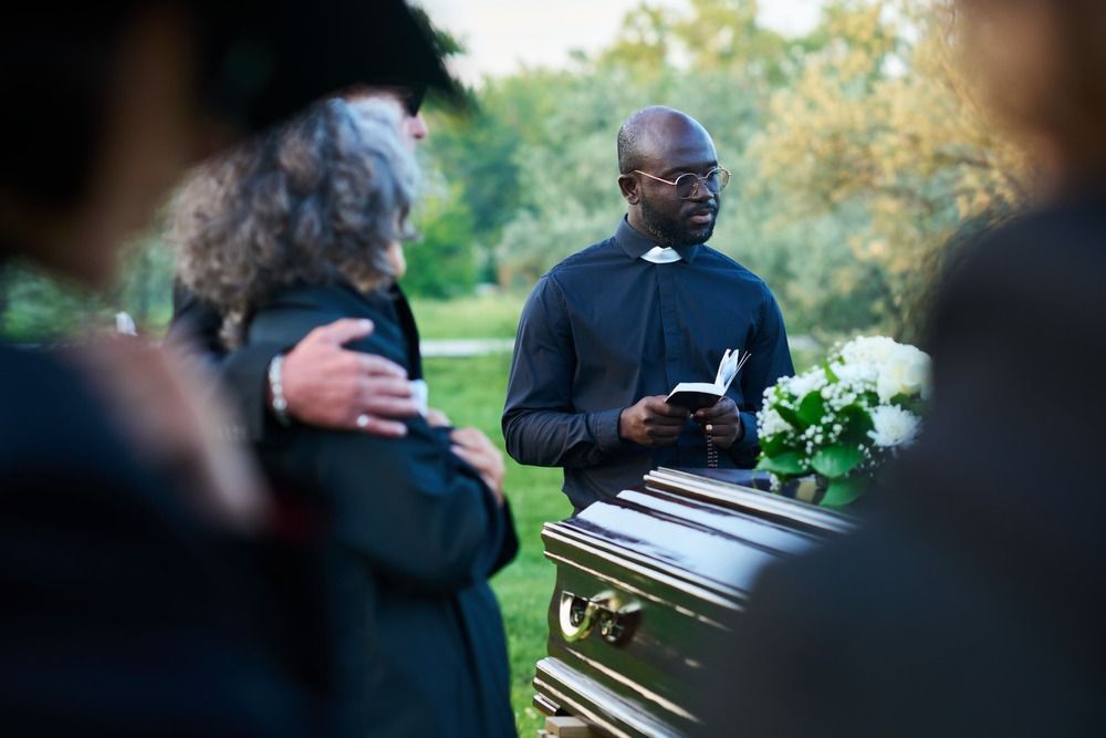 Mourners stand beside a casket as a clergy member holds a book at an outdoor funeral service.