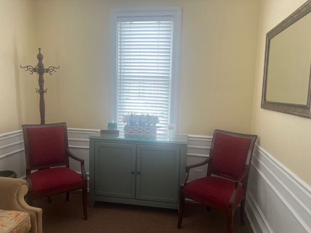 Small waiting room with two red chairs, a green cabinet, wall mirror, and coat rack by a window.