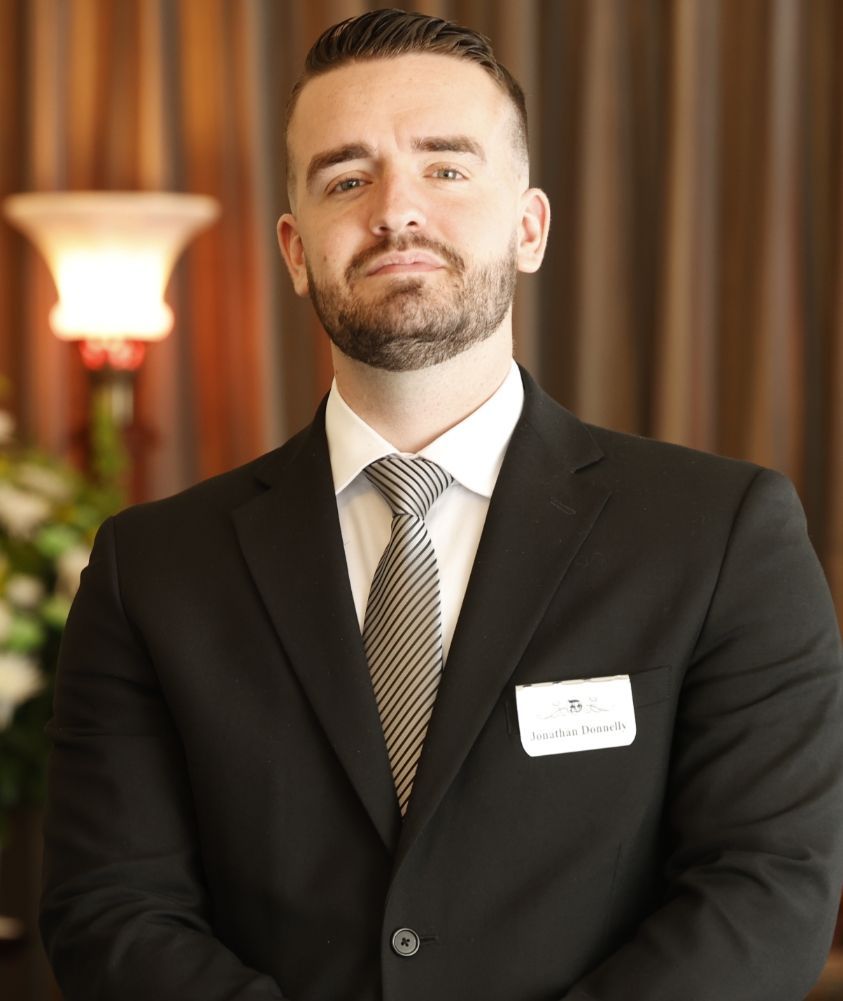 Man in a black suit and tie with a name badge, standing indoors against a blurred brown backdrop.