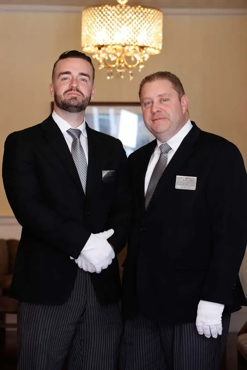 Two formally dressed people stand side by side in a chandelier-lit room, wearing white gloves.