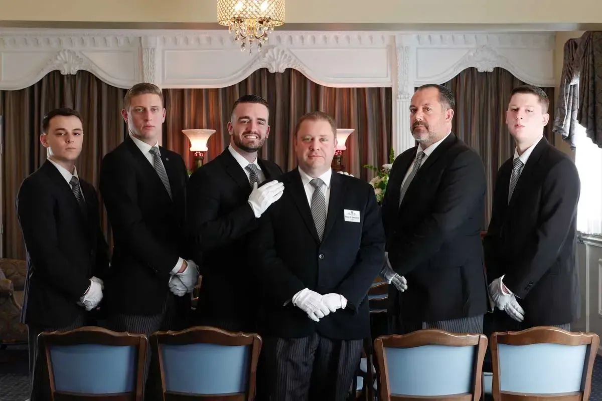 Five men in tuxedos and white gloves pose in a formal dining room with chandeliers and chairs