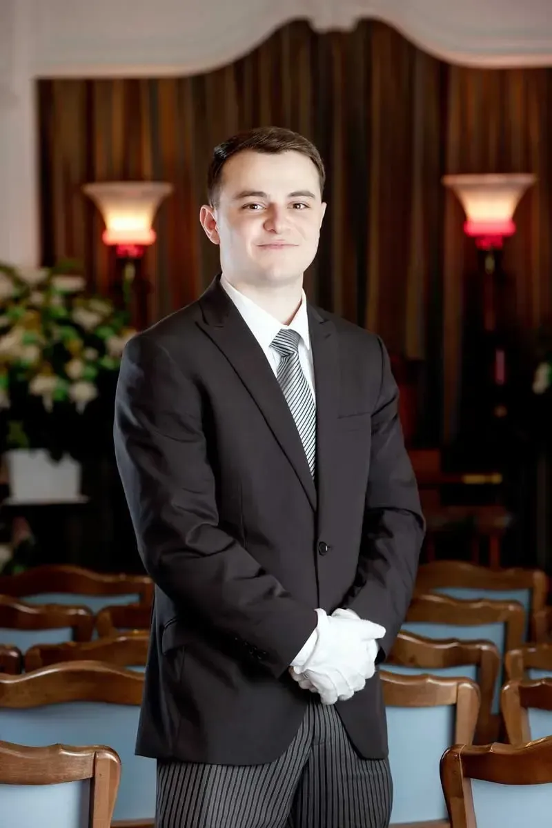 Person in a black suit and white gloves standing in a formal room with rows of chairs and warm wall lamps.