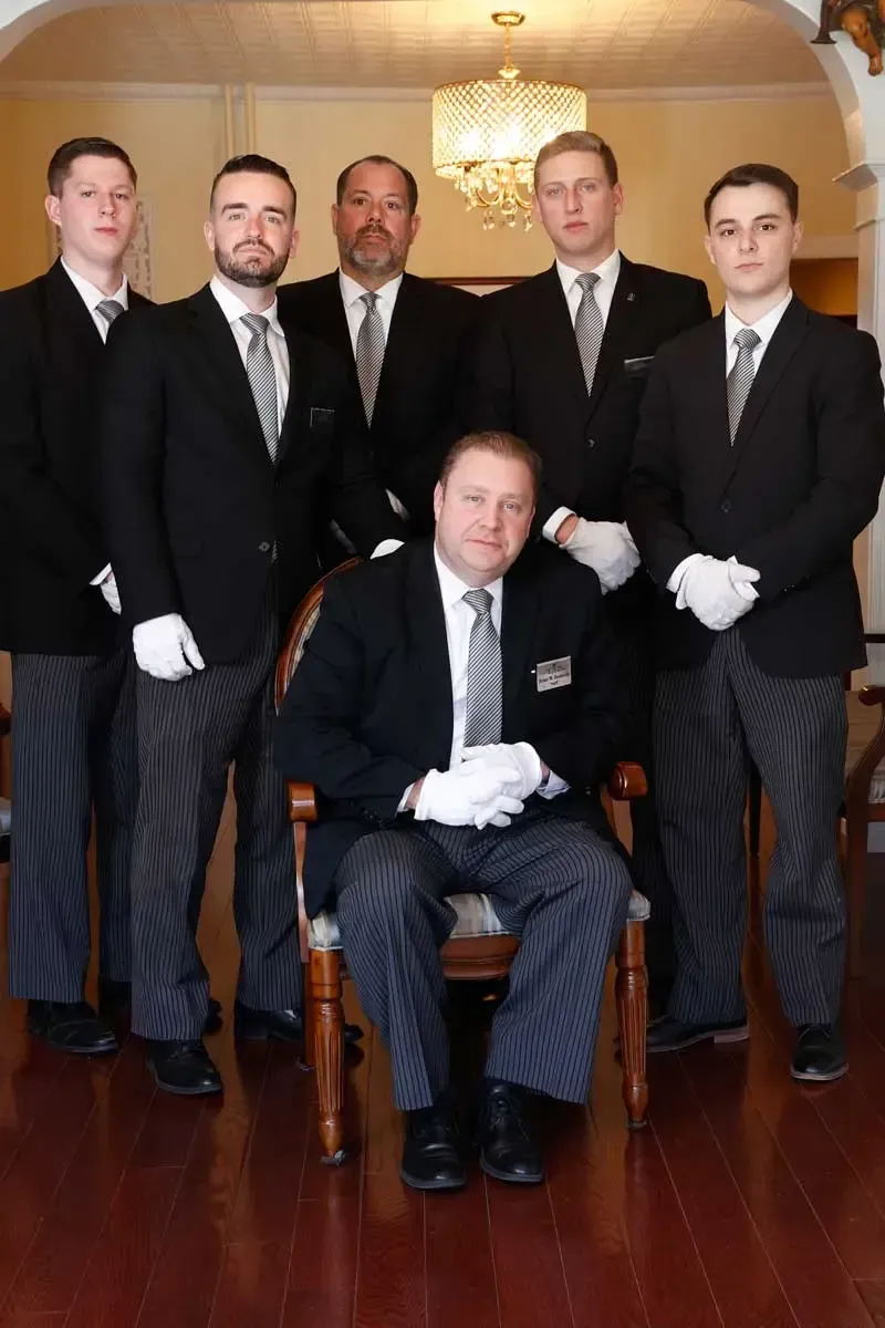 Six men in dark suits pose indoors, four standing and one seated in front, in a formal room.
