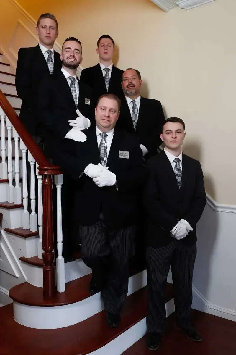 Five men in black suits and white gloves pose on a staircase in a hallway