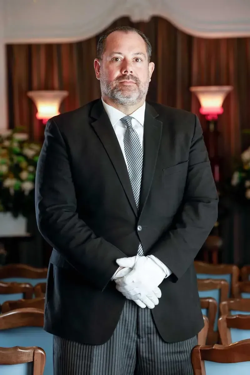 A man in a black suit and white gloves stands indoors between rows of chairs.