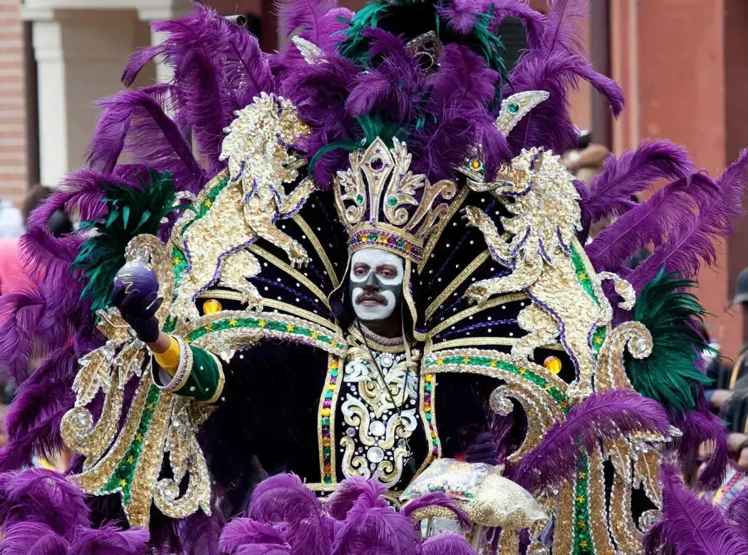 Masked performer in a purple feathered carnival costume with a jeweled headdress and white face paint