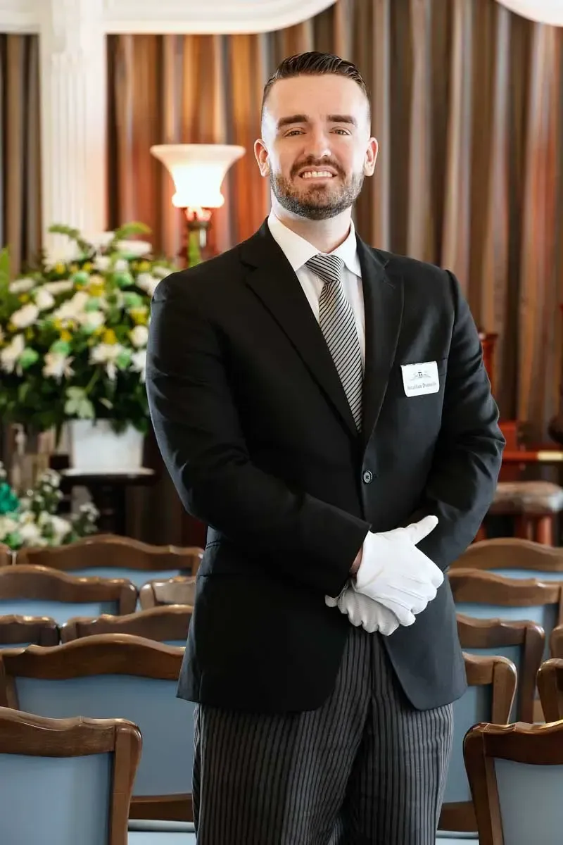 Man in a black suit and gloves stands smiling in a formal room with chairs and flowers.