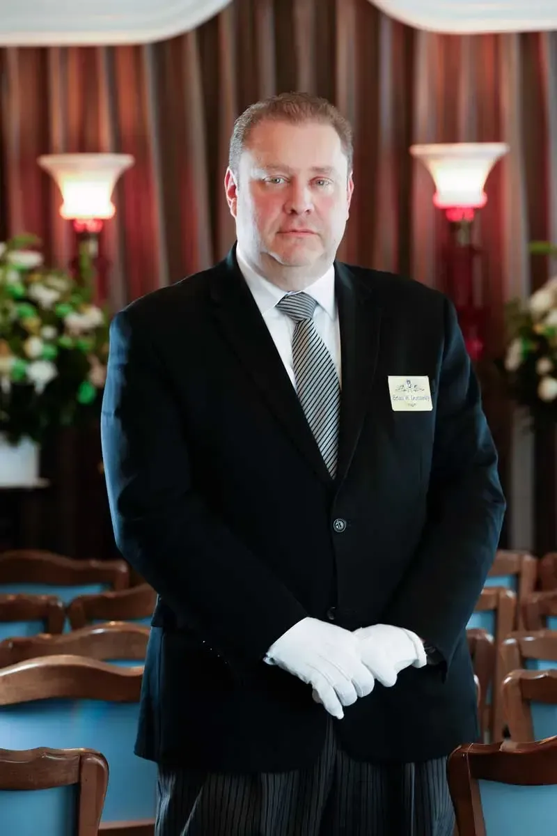 Man in a black suit and white gloves standing in a formal event room with chairs and red curtains.