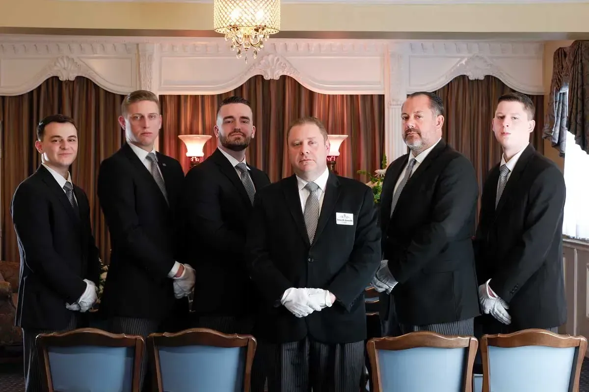Six men in dark suits standing in a formal room with draped windows and a chandelier