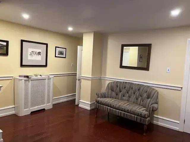 Empty waiting room with beige walls, framed art, white cabinet, and a striped loveseat on wood floors